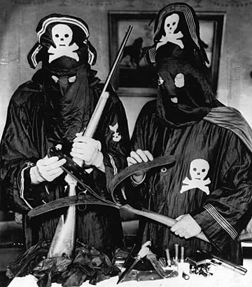 In this 1936 image from the photo agency, Acme News Photos (ACME), Detroit police officers pose with weapons and regalia seized from Black Legion terrorists. The officers, dressed in the black robes and pirate hats of the Legionnaires, display a captured lever-action rifle, a .45 caliber 1911 pistol, and a leather whip used to flog victims. The image was an ACME "press photo" circulated to various news publications in '36. Photographer unknown.