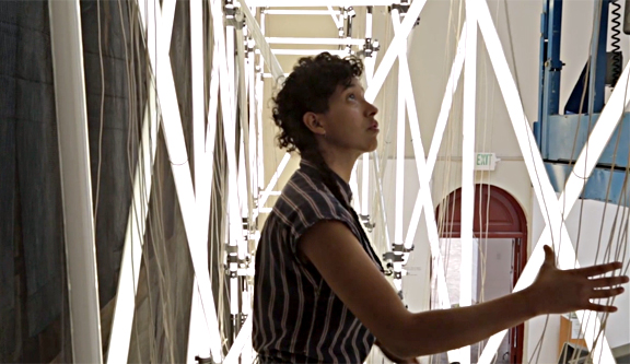 Pereda's installation of scaffolding and fluorescent lighting is inches away from Diego Rivera's mural, hidden on the left by black construction netting. In this Screen grab from the SFIA short film, Change the World or Go Home, an assistant of Pereda's adjusts the fluorescent lights.
